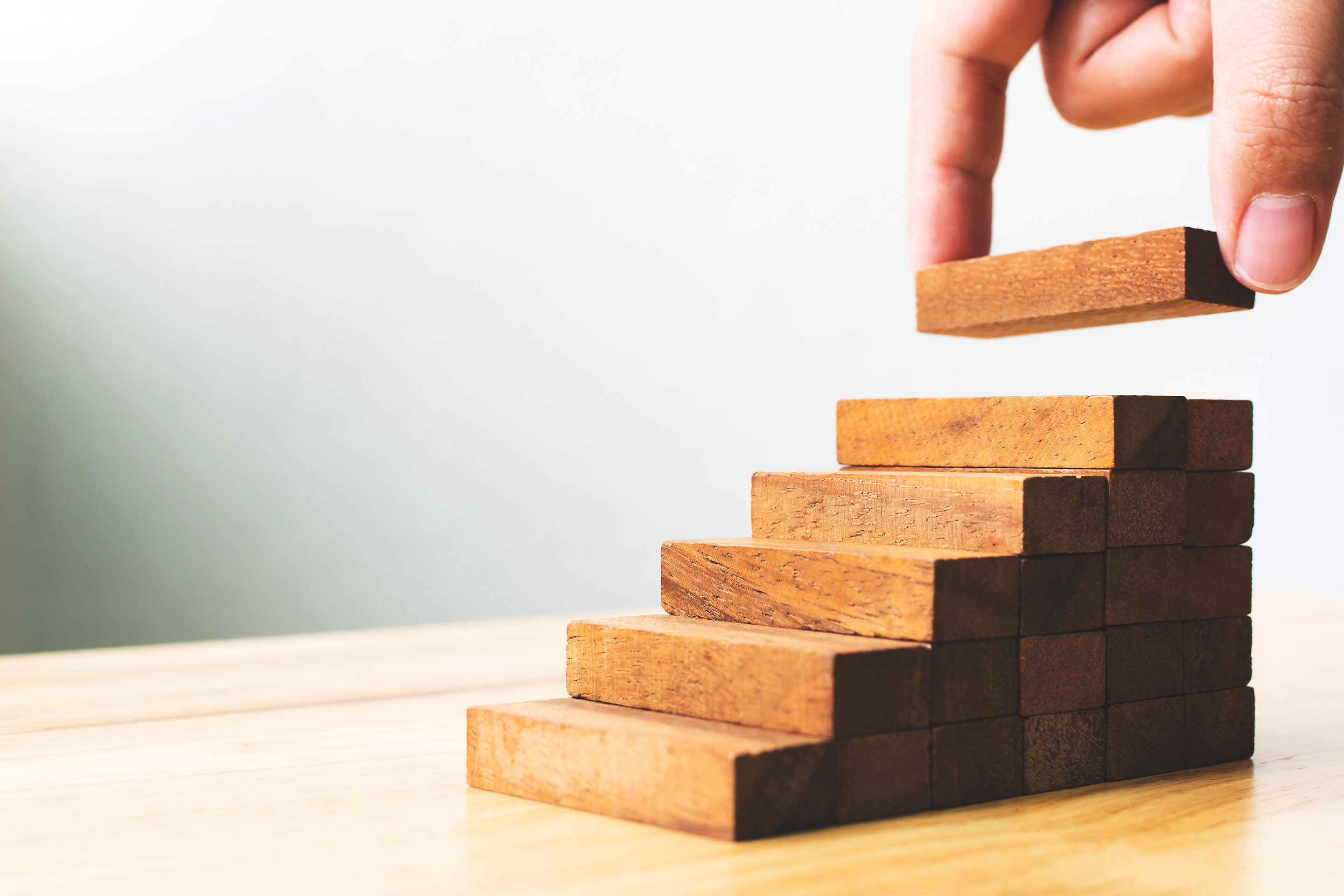 A hand adds a block to a wood puzzle in the shape of ascending stairs, as a metaphor for potential investment performance