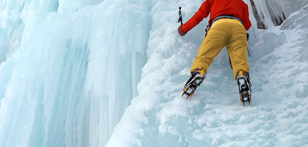 A clinber in red and yellow snow gear scales a steep cliff covered in ice. He's supported by climbing gear and boots.