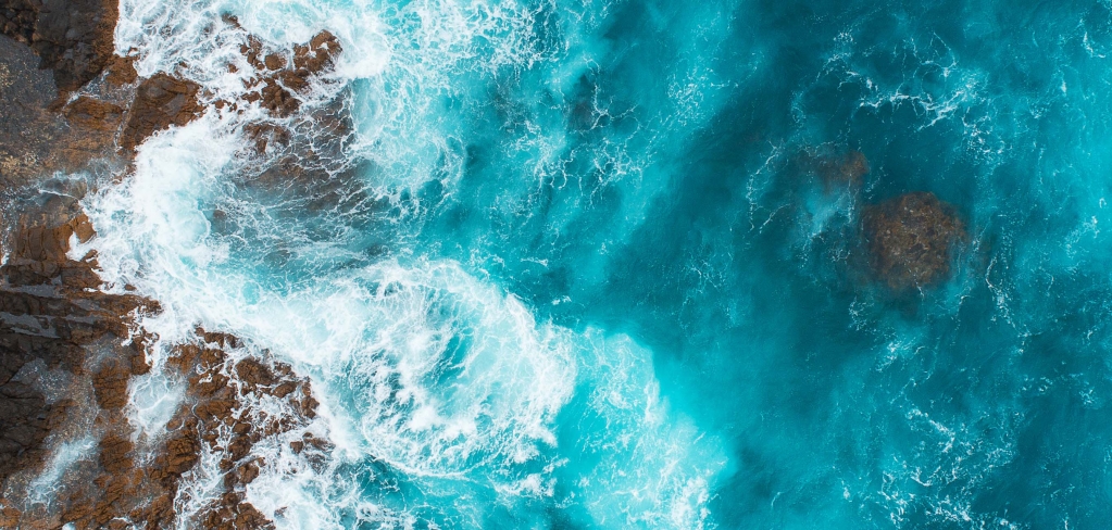 Aerial view of bright blue ocean water crashing into the side of a cliff.