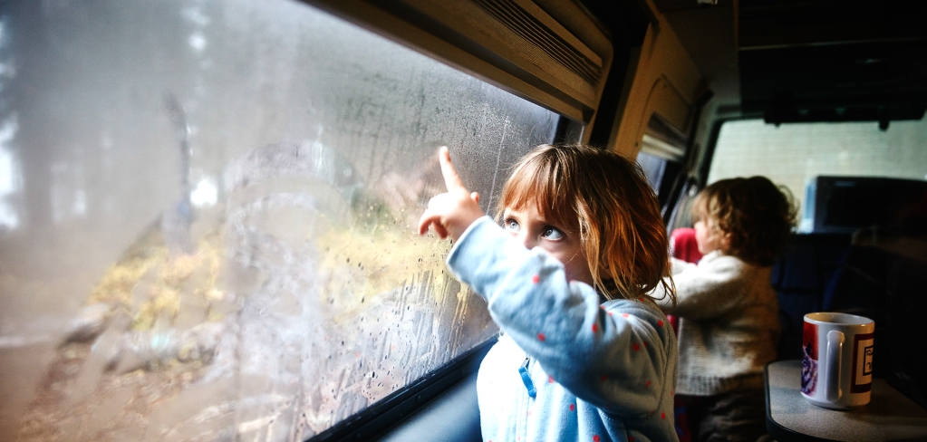 Family vacation: Two small children watch water droplets on the windows of a van, which is traveling through a forest.